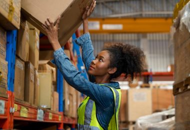 A worker wearing a yellow safety vest lifting a cardboard box from a shelf in a warehouse aisle with multiple racks.