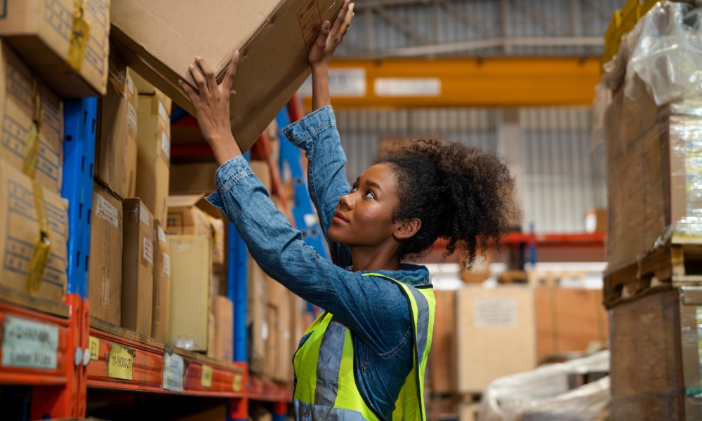 A worker wearing a yellow safety vest lifting a cardboard box from a shelf in a warehouse aisle with multiple racks.