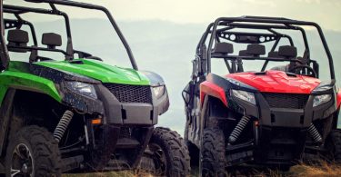 A green off-road vehicle and a red off-road vehicle parked in dry grass with cloudy sky and mountains behind them.