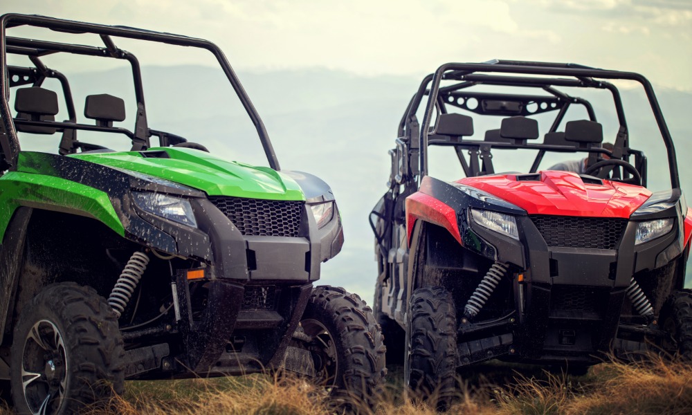 A green off-road vehicle and a red off-road vehicle parked in dry grass with cloudy sky and mountains behind them.