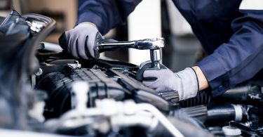 A person wearing gray gloves using a socket wrench on an open car engine inside an auto repair shop.