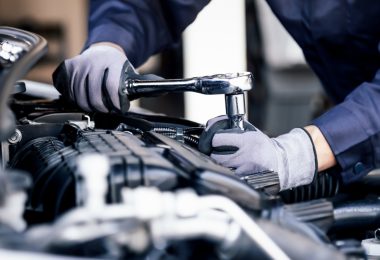 A person wearing gray gloves using a socket wrench on an open car engine inside an auto repair shop.