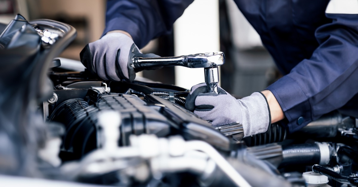 A person wearing gray gloves using a socket wrench on an open car engine inside an auto repair shop.