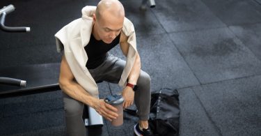 A man sitting on a gym bench while holding a half-full shaker bottle, with a towel draped over his shoulders.