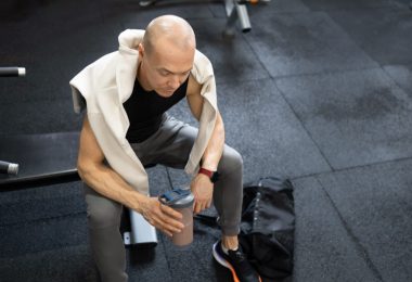 A man sitting on a gym bench while holding a half-full shaker bottle, with a towel draped over his shoulders.