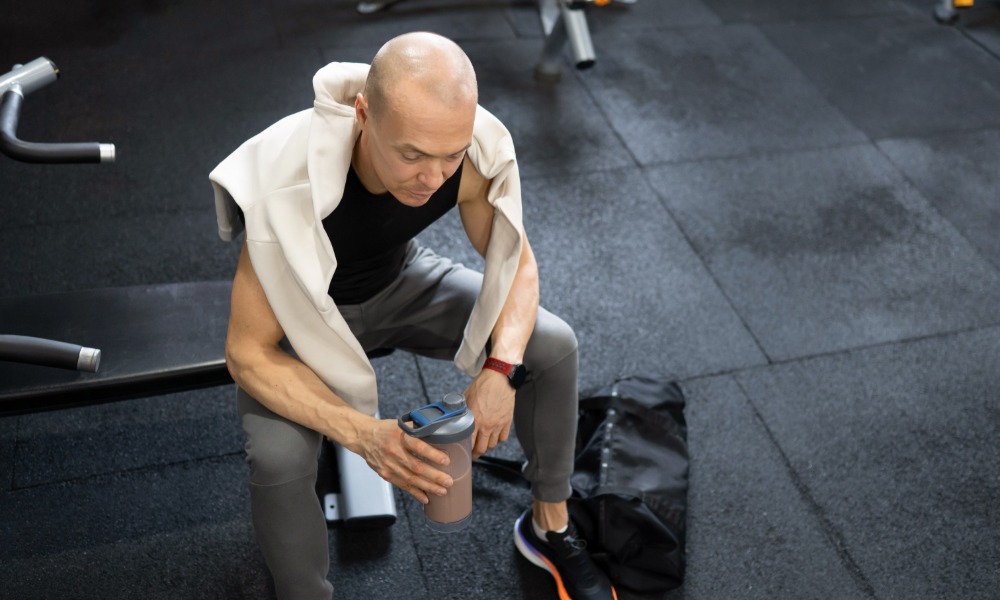 A man sitting on a gym bench while holding a half-full shaker bottle, with a towel draped over his shoulders.