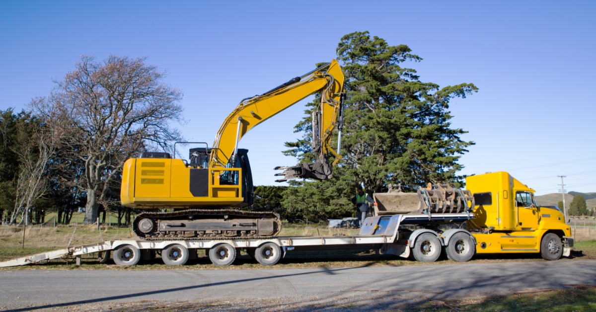 A yellow excavator secured on a step deck trailer attached to a yellow semi-truck parked on a rural roadside.