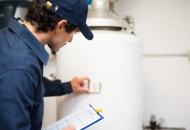 A plumber standing beside a white water heater while examining it with his hand. He is also holding a clipboard.
