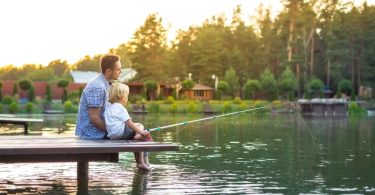 A father and son sitting on a dock with a fishing line in the water. There are lake houses behind them.