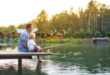 A father and son sitting on a dock with a fishing line in the water. There are lake houses behind them.