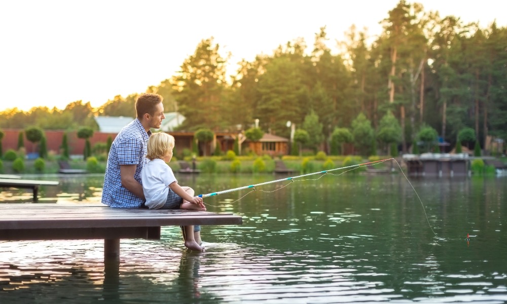 A father and son sitting on a dock with a fishing line in the water. There are lake houses behind them.