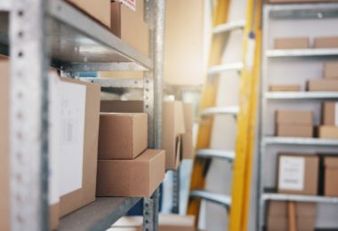A storage room with cardboard boxes on metal shelves and a yellow ladder standing between narrow aisles.