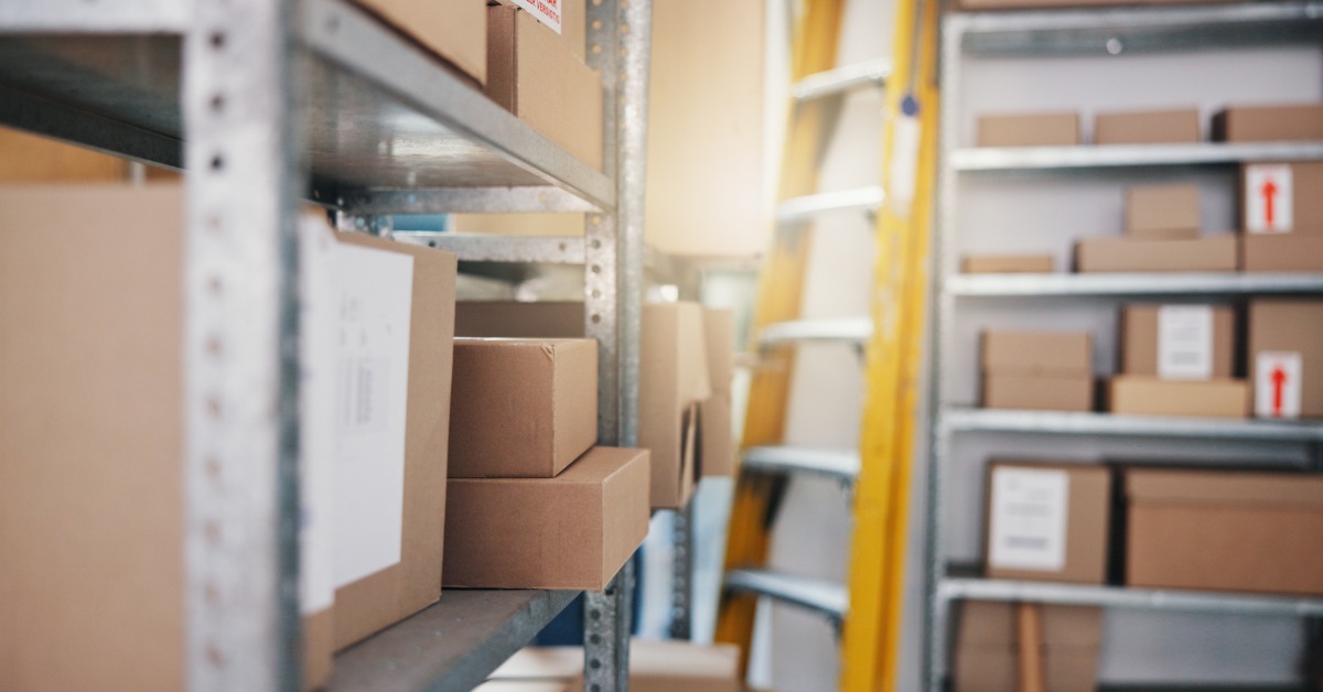 A storage room with cardboard boxes on metal shelves and a yellow ladder standing between narrow aisles.
