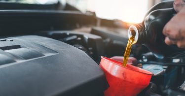A close-up of a person pouring engine oil from a bottle into a red funnel. Sunlight shines into the garage in the background.