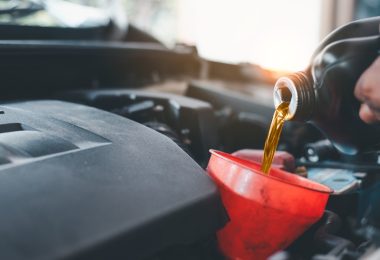 A close-up of a person pouring engine oil from a bottle into a red funnel. Sunlight shines into the garage in the background.