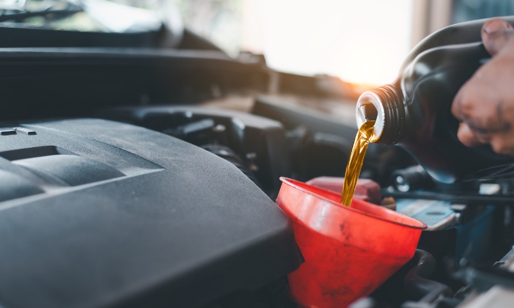 A close-up of a person pouring engine oil from a bottle into a red funnel. Sunlight shines into the garage in the background.