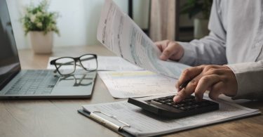 A close-up of a person reviewing financial paperwork and using a calculator at a desk containing a laptop and glasses.