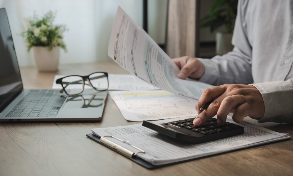 A close-up of a person reviewing financial paperwork and using a calculator at a desk containing a laptop and glasses.