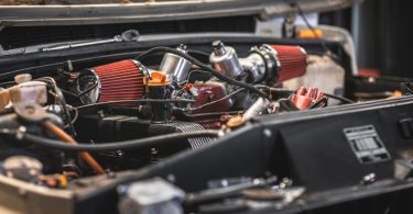 A car engine compartment with red air filters, metal parts, black panels, hoses, wires, and a raised hood.