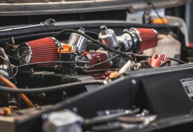 A car engine compartment with red air filters, metal parts, black panels, hoses, wires, and a raised hood.
