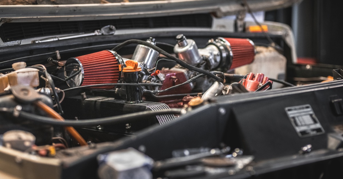 A car engine compartment with red air filters, metal parts, black panels, hoses, wires, and a raised hood.