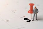 A miniature businessman figure standing beside a red pushpin and black dollar sign on a paper calendar.