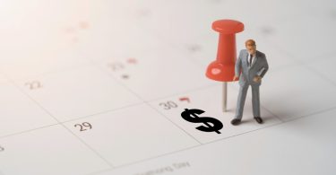 A miniature businessman figure standing beside a red pushpin and black dollar sign on a paper calendar.