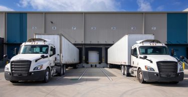 Two white semi-trucks with trailers parked outside numbered loading docks at a large warehouse under a blue sky.