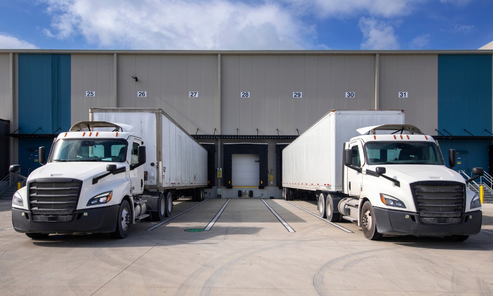 Two white semi-trucks with trailers parked outside numbered loading docks at a large warehouse under a blue sky.
