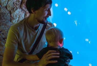 A father holding his young child as they look into a brightly lit blue aquarium filled with jellyfish.