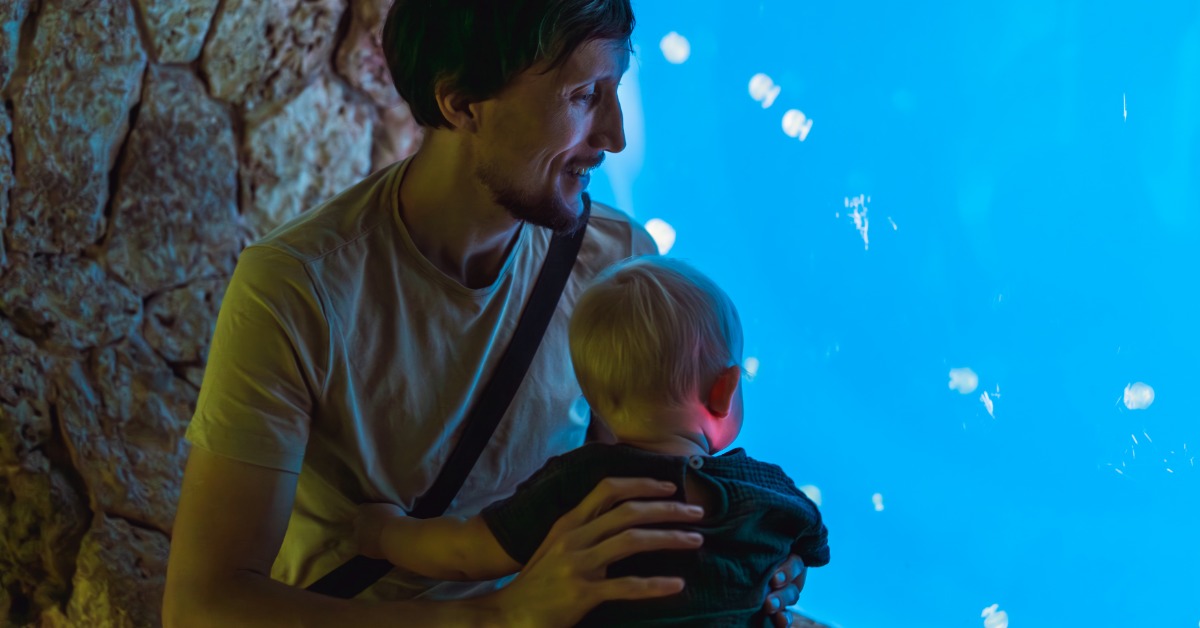 A father holding his young child as they look into a brightly lit blue aquarium filled with jellyfish.