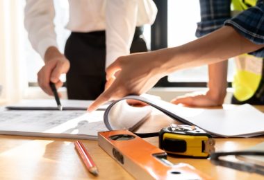 A hand points to papers on a table beside an orange level, yellow tape measure, pencil, helmet, and gloves.