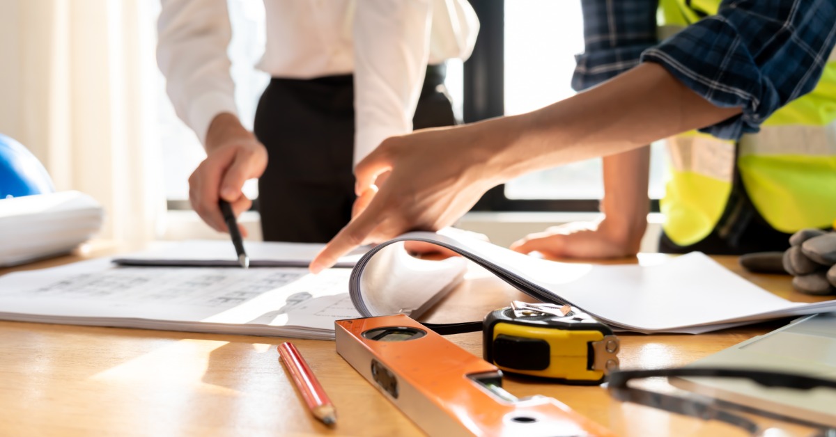 A hand points to papers on a table beside an orange level, yellow tape measure, pencil, helmet, and gloves.