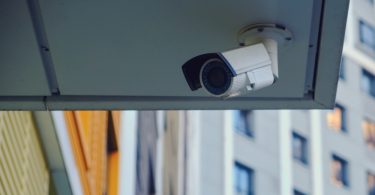 White security camera mounted beneath an apartment building overhang with neighboring apartments visible behind it.