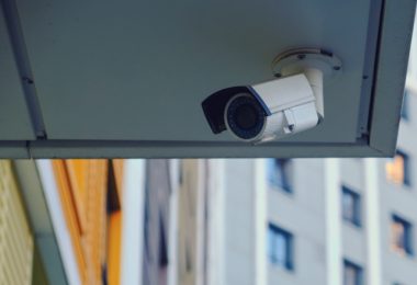 White security camera mounted beneath an apartment building overhang with neighboring apartments visible behind it.