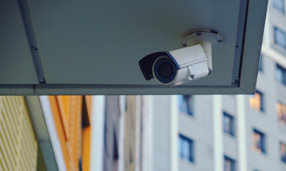 White security camera mounted beneath an apartment building overhang with neighboring apartments visible behind it.