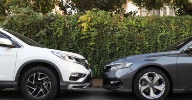 A low-angle view of a white car and a gray car parked nose to nose on pavement in front of bushes and trees.