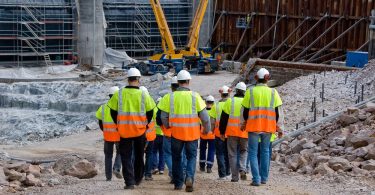 Construction workers in high-visibility vests and hard hats walking together through a large active jobsite.