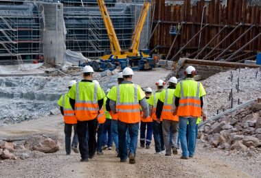 Construction workers in high-visibility vests and hard hats walking together through a large active jobsite.