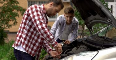 A man in a red plaid shirt tightens a component beneath the hood of a car. A teenager stands and watches nearby.