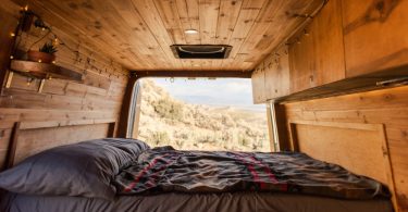 A camper van interior with a bed, grey bedding, wood cabinets, a small succulent, and the rear door open.