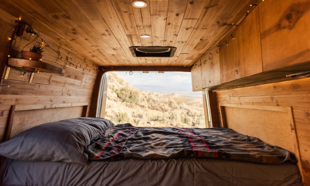 A camper van interior with a bed, grey bedding, wood cabinets, a small succulent, and the rear door open.