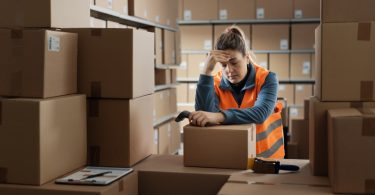 A person in a safety vest leaning on stacked cardboard boxes in a warehouse aisle, with one hand touching their head.