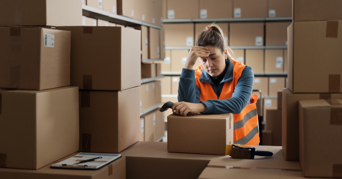 A person in a safety vest leaning on stacked cardboard boxes in a warehouse aisle, with one hand touching their head.