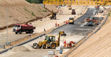 Multiple workers operating heavy equipment and trucks to build a roadway with fresh pavement and curbs.
