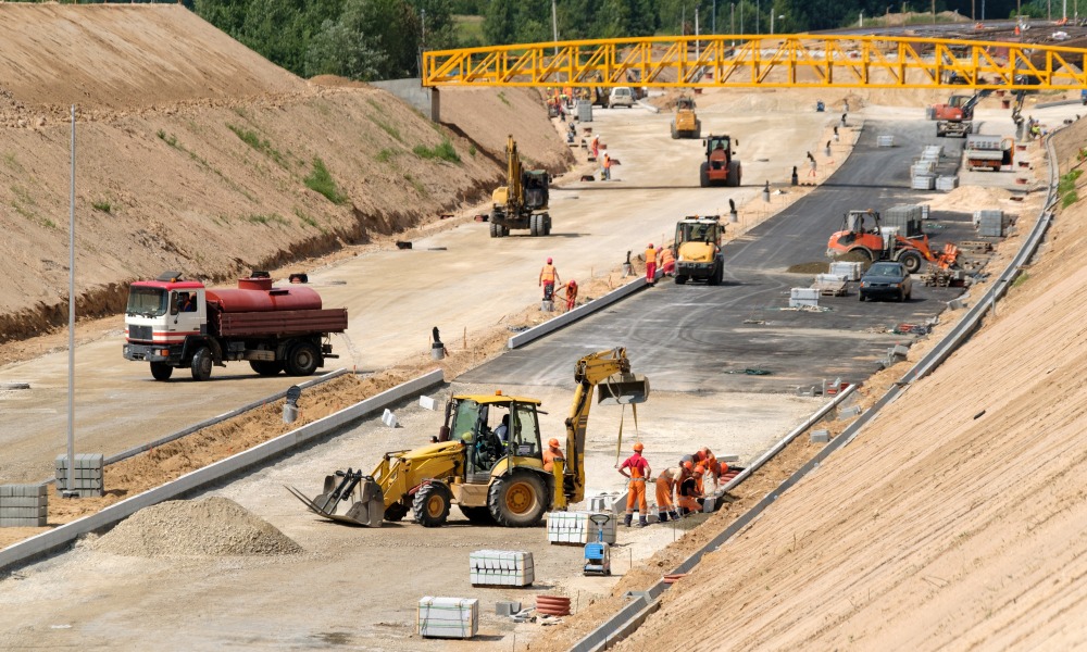 Multiple workers operating heavy equipment and trucks to build a roadway with fresh pavement and curbs.
