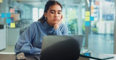 Tired woman rests her face on her hand while looking at a laptop at her desk in a bright modern office.