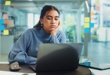 Tired woman rests her face on her hand while looking at a laptop at her desk in a bright modern office.