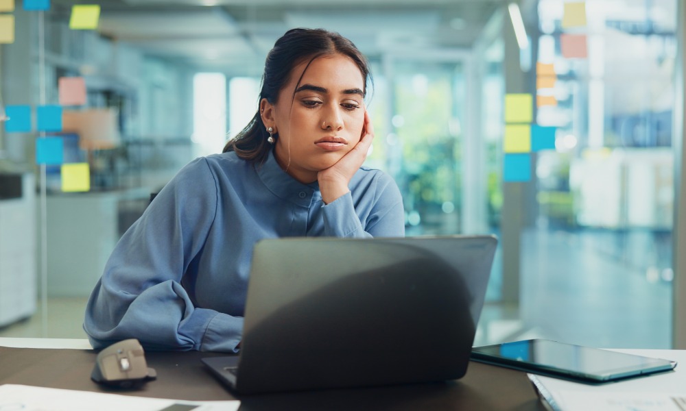 Tired woman rests her face on her hand while looking at a laptop at her desk in a bright modern office.