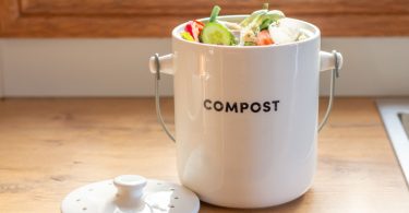 A white compost container filled with vegetable scraps sits on a wooden kitchen counter beside its lid.